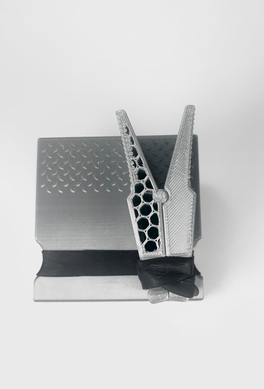 A metallic silver 3D-printed object with a textured honeycomb pattern on one side and a lattice design on the other, resting on a black rectangular base against a light gray background.